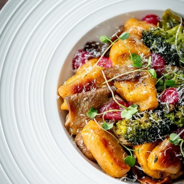 Close-up of golden gnocchi with broccoli, mushrooms, red berries, and microgreens in a white bowl.