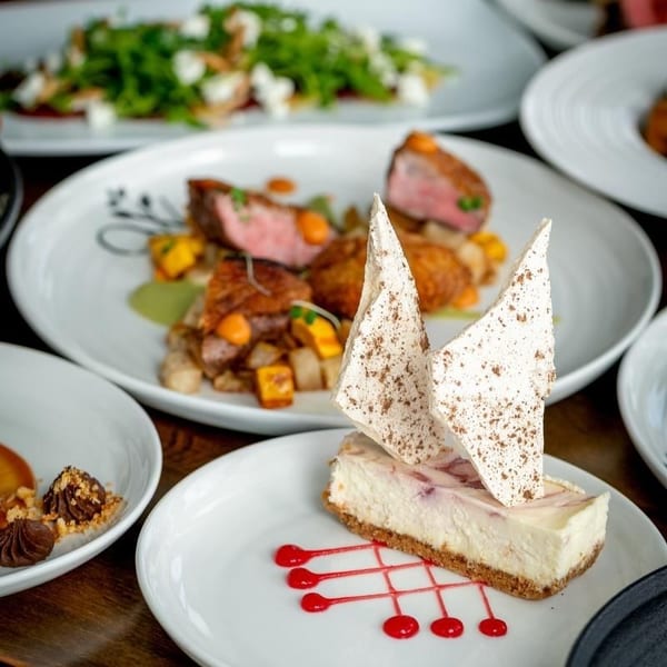 A slice of cheesecake with meringue shards and raspberry drizzle, with steak and salad in the background.